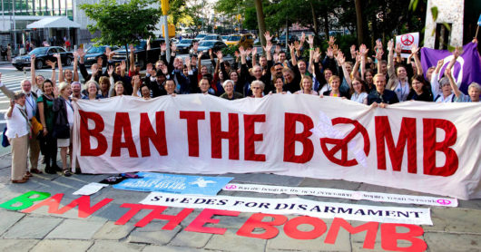 Joukko ydinasekieltoa kannattavia aktivisteja hurraa ja pitelee valkoista banderollia, jossa lukee punaisin kirjaimin "Ban the Bomb"