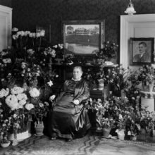 Lucina Hagman istuu tuolissa huoneessa, ympärillä on kukkia.