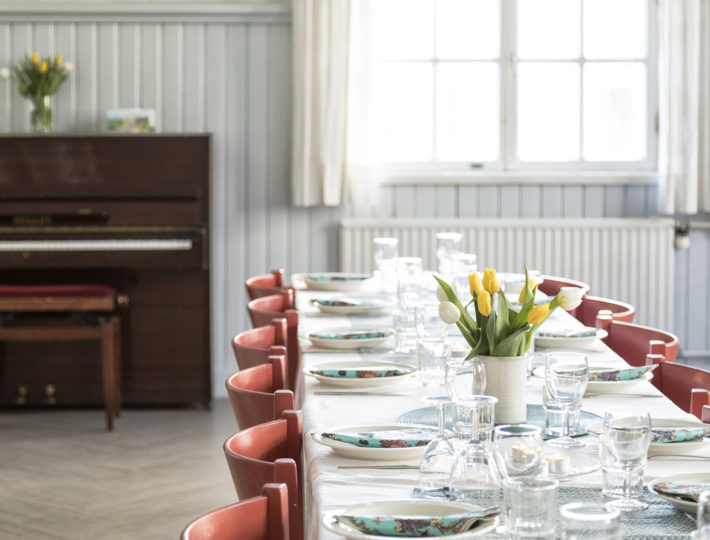 Kuvassa Rauhanaseman juhlasaliin on katettu pitkä pöytä. Pöydällä on valkoinen pöytäliina, tulppaaneja ja astiat. Taustalla näkyy ikkuna ja piano, jonka päällä on keltaisia kukkia.